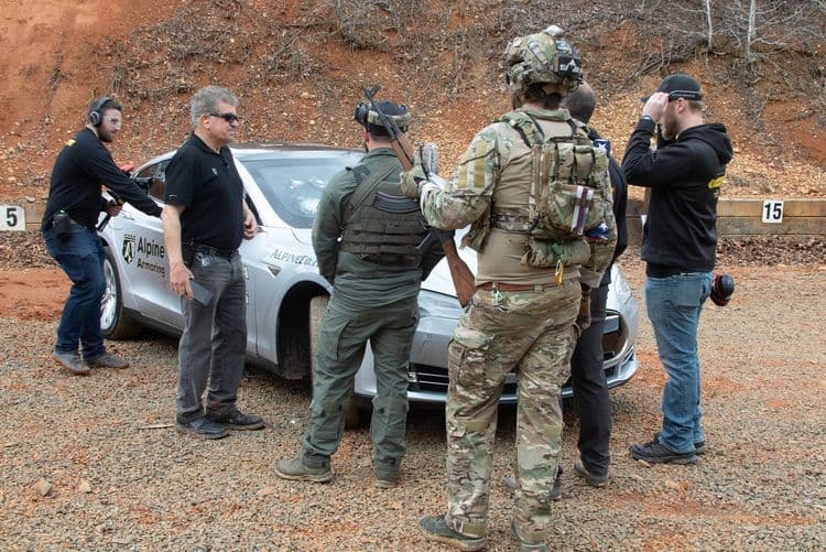 A group of men in tactical gear and headphones gather around a parked car in an outdoor setting.