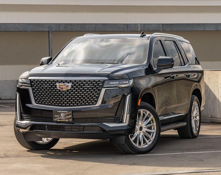 A black armored Cadillac Escalade Premium Luxury with a Cadillac emblem parked in a concrete parking lot, featuring large wheels and a prominent front grille.