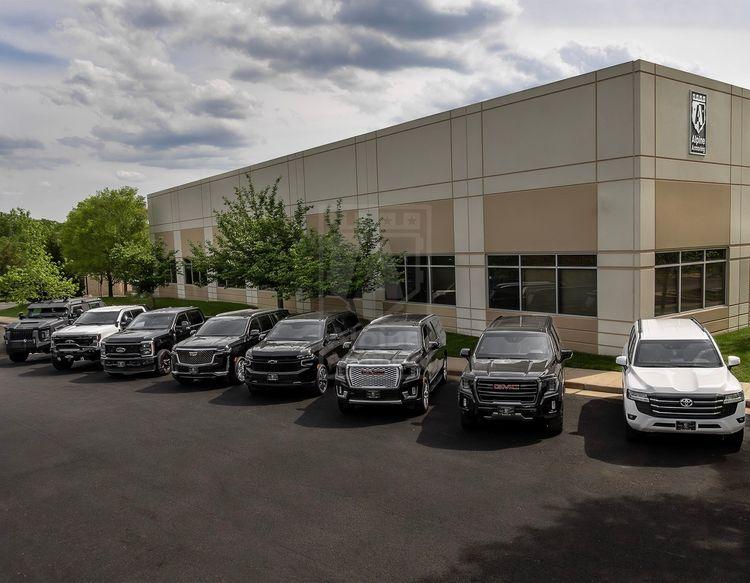 A lineup of various black and one white armored vehicles parked in front of the Alpine Armoring facility. The building features the Alpine Armoring logo, and the setting includes a well-maintained landscape with trees and a partly cloudy sky.