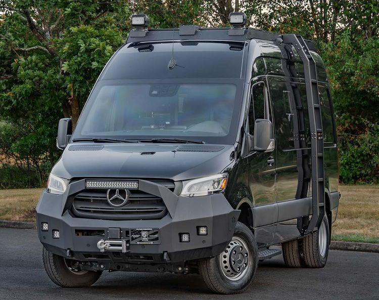 A modified black Mercedes-Benz Pointer van from Alpine Armoring with rugged features including additional lights, a winch, and reinforced bumpers. It's parked on a paved road with trees in the background.