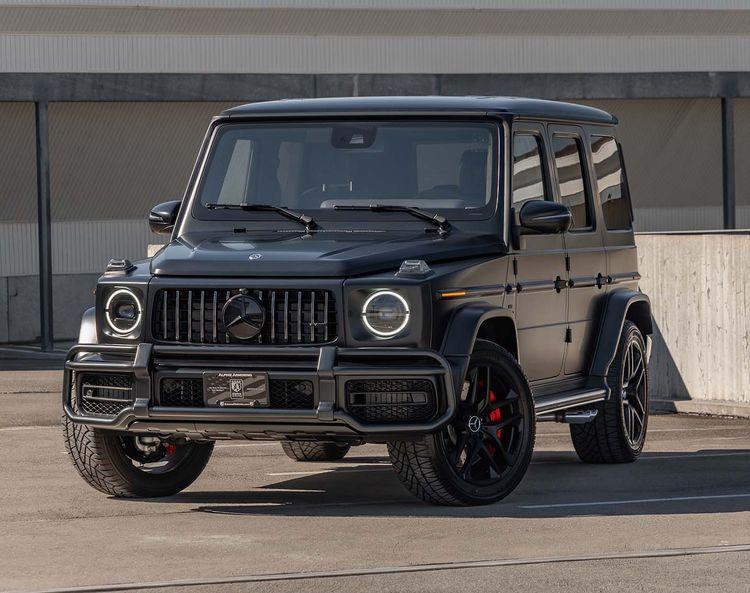 A black armored Mercedes-Benz G63 AMG SUV with distinctive round headlights is parked in an industrial-looking area on a sunny day.