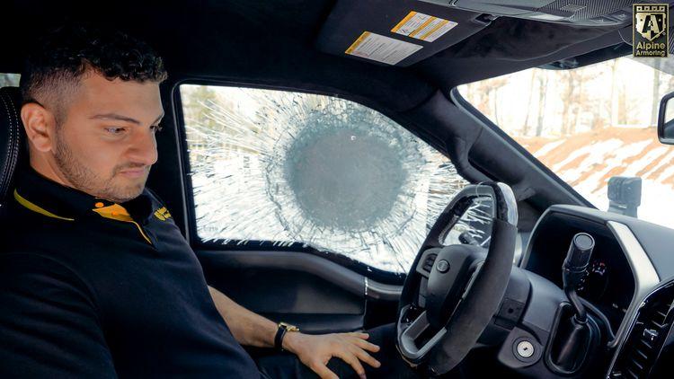 A man sits in the driver's seat of an Alpine Armoring MASTIFF with a severely cracked windshield. He is wearing a black shirt with a yellow collar. The windshield has a large impact mark on the driver's side. The logo in the image says "Alpine Armoring.