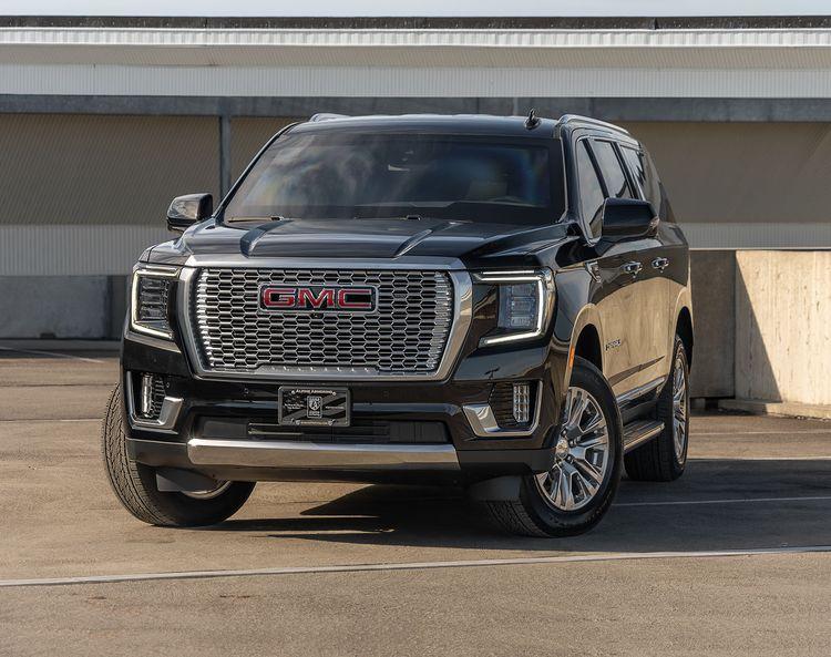 Front view from the left side of a black armored GMC Yukon Denali SUV with dual exhaust pipes, displayed in a parking area. The vehicle has "Alpine Armoring" branding on the back.