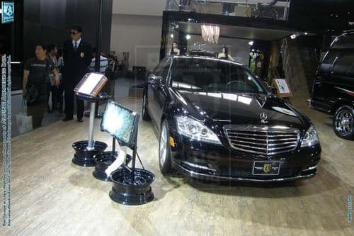 A black luxury sedan is displayed indoors at an auto show. The vehicle has a prominent Mercedes emblem on the front grille. Display stands with information and digital screens are positioned around the car. People can be seen in the background.