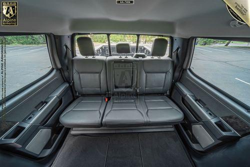 A rear interior view of a bulletproof Ford F-350 Lariat showcasing three grey leather seats, headrests, and a spacious legroom area with black floor mats. The background shows an empty parking lot.