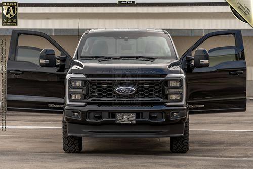 Front view of an armored black Ford F-350 Lariat truck parked in an outdoor lot. The truck features large side mirrors, a prominent grille, and is positioned against a concrete background with overhang.