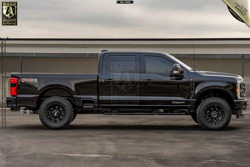 Passenger side view of a black bulletproof Ford Super Duty F-350 Lariat pickup truck parked on a concrete surface in front of a building. The truck has large off-road tires and a crew cab configuration.