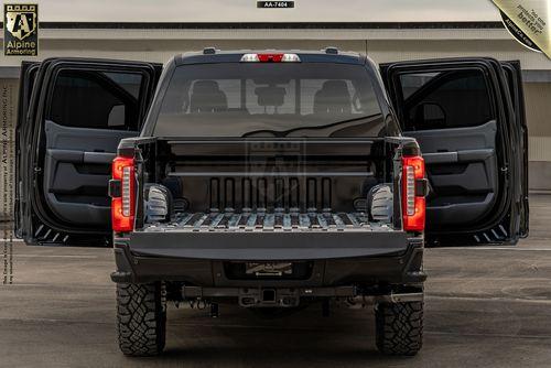 A black armored Ford F-350 Lariat truck with its tailgate and rear doors open, showcasing the empty truck bed and vehicle's interior. Labeled "Alpine Armoring" with a product number "AJAX7044" at the top.