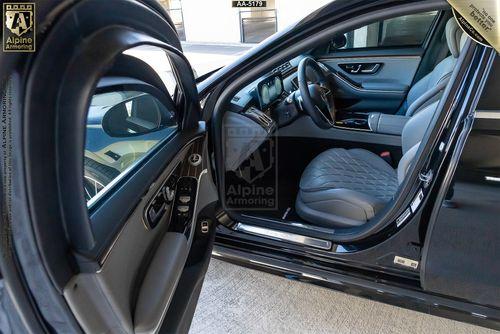 Interior view of a black armored Mercedes-Benz S580 S-Class 4MATIC showing the driver's seat, passenger seat, dashboard, and door with visible branding for Alpine Armoring.