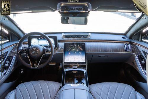 Interior of a black armored Mercedes-Benz S580 S-Class 4MATIC sedan showing the dashboard, steering wheel, large central touchscreen, quilted leather seats, and wood trim accents. The badge on the dashboard indicates Alpine Armoring.