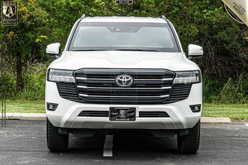 A white Armored Toyota Land Cruiser 300 SUV is parked in a parking lot with trees in the background. The front grille and headlights are visible.