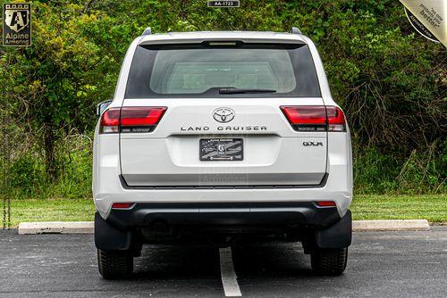 Rear view of a white armored Toyota Land Cruiser 300 SUV parked in a lot with trees and foliage in the background. License plate area blurred.