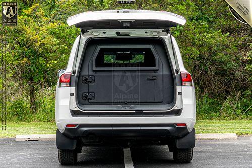 A white Armored Toyota Land Cruiser 300 SUV with the rear hatch open, revealing a secure storage compartment in the back. Trees and grass are visible in the background.