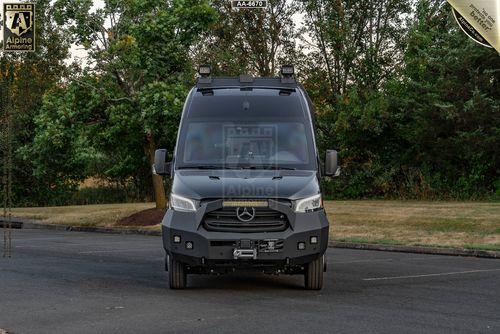 A black armored SWAT Pointer Van from Alpine Armoring with a Mercedes-Benz logo is parked on a paved road with trees in the background.