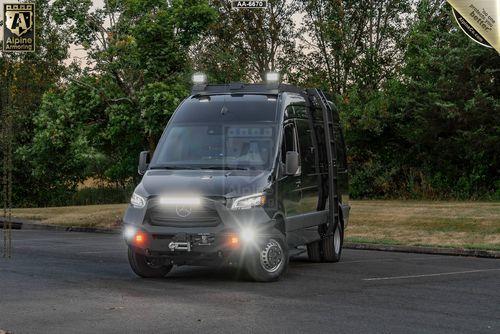 A black armored SWAT Pointer Van from Alpine Armoring with bright headlights is parked in an outdoor area with trees in the background. The vehicle displays the Alpine Armoring logo and has various equipment mounted on the roof.