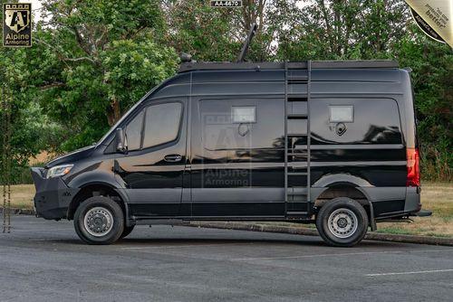 A black armored SWAT Pointer Van from Alpine Armoring is parked at the side of a street. The van has tinted windows, a rooftop railing, and a side ladder. Trees and grass are visible in the background.