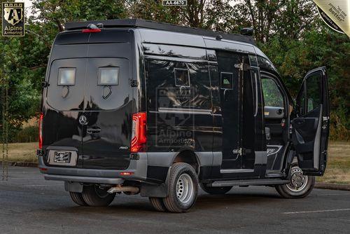 A black, armored SWAT Pointer Van from Alpine Armoring with doors open, parked outdoors. The van displays Alpine Armoring logos and has several windows. Trees are visible in the background.