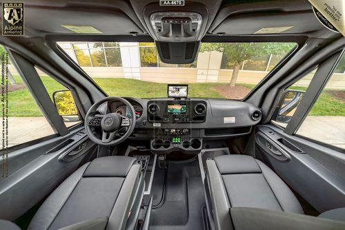 The interior of a SWAT Pointer Van from Alpine Armoring, showing the driver's seat, steering wheel, dashboard with digital controls, and two front seats. The vehicle has large windows and a view of an outside building and trees.