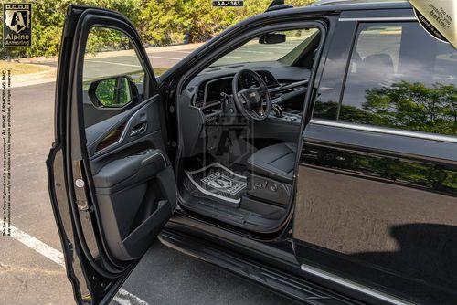 A bulletproof black Cadillac Escalade ESV Luxury SUV with an open driver's side door showcasing the interior, including the steering wheel, dashboard, and driver's seat. The vehicle is parked on an asphalt surface near green foliage.