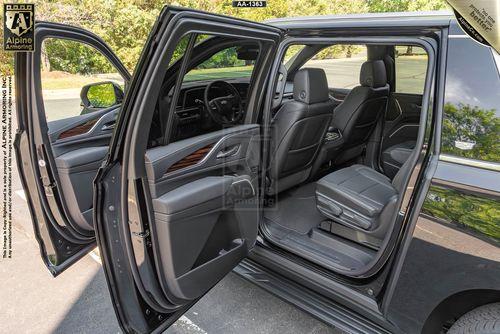 A black bulletproof Cadillac Escalade ESV Luxury SUV with all doors open, showcasing a spacious and luxurious interior with black leather seats. The vehicle is parked in a sunny outdoor setting with trees in the background.