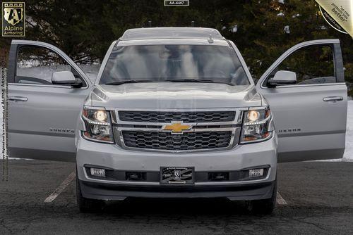 A front view of a white Chevrolet Suburban 3500HD LT VIP Limo with its front doors open in a parking lot. Snow and trees are visible in the background. Alpine Armoring logos are displayed in the corners.