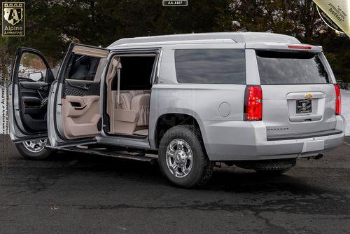 A silver bulletproof  Chevrolet Suburban 3500HD LT VIP Limo with its rear doors open, showing a beige interior and additional features such as side steps and a spare tire mounted below. The vehicle is parked on a paved surface outdoors.