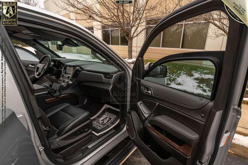 Open doors of a bulletproof Chevrolet Suburban 3500HD LT VIP Limo revealing a black leather interior with a dashboard, steering wheel, and a white mat on the floor. Outside, snow is visible near a beige building.