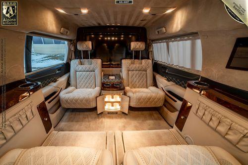 Luxurious Chevrolet Suburban 3500HD LT VIP Limo interior featuring plush, white leather seating, wood paneling, and ambient lighting. The setup includes a table, cup holders, and a large screen at the front.