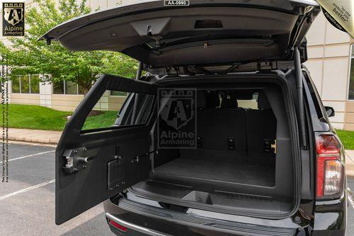 An open rear view of a black armored Chevrolet Suburban LT  SUV showing its empty cargo area and a transparent cover with the Alpine Armoring logo. Trees and a building are in the background.