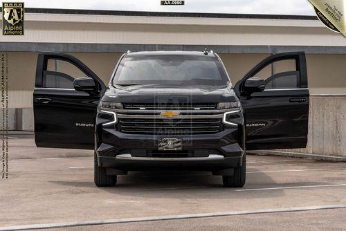 A black bulletproof Chevrolet Suburban LT is parked with its front doors open, facing the camera. The backdrop includes a concrete parking area and a building.