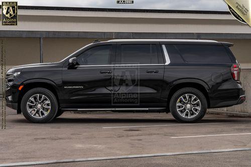 A black armored Chevrolet Suburban LT SUV labeled "Suburban" is parked in a lot near a building, displaying a logo from Alpine Armoring.