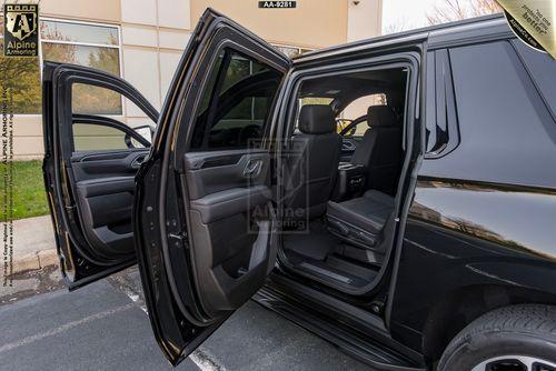 A black, bulletproof Chevrolet Suburban RST SUV with open front and rear passenger doors parked on a street in front of a building.