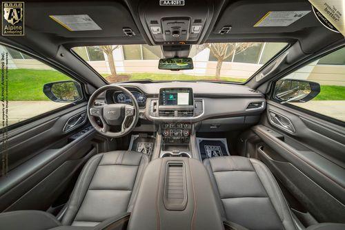 Driver's seat perspective view of a Chevrolet Suburban RST dashboard with a central touchscreen display, steering wheel with controls, and black leather seats. Through the windshield, trees and a building are visible.