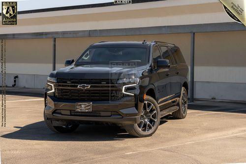 A black armored Chevrolet Suburban RST SUV is parked in an industrial setting on a sunny day.