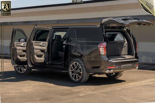 A black armored Chevrolet Suburban RST SUV is parked with all doors, including both rear and tailgate doors, open. The vehicle is in an outdoor setting with a beige building wall in the background.