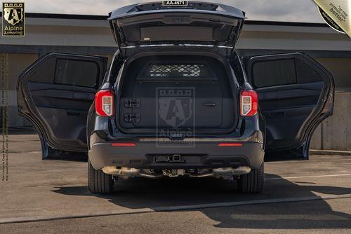 The rear view of a black Ford Explorer Police Protection Vehicle (PPV) SUV is shown with its rear door, tailgate, and rear passenger doors open, revealing the cargo area. The vehicle is parked on a concrete surface near a building.