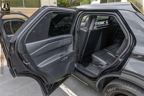 The open rear door of a Ford Explorer Police Protection Vehicle revealing the interior. The back seat is empty and several windows are partially visible. The vehicle is parked in a lot next to a beige building.