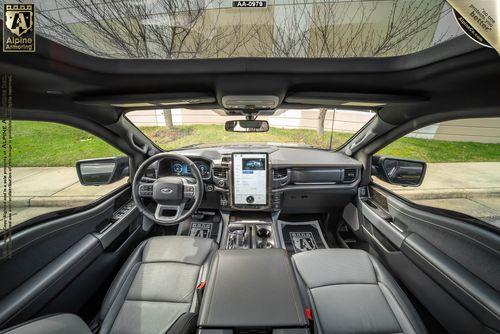 Interior view of a Ford Lightning showcasing its dashboard, steering wheel, large central touchscreen display, and panoramic sunroof. The logo "Alpine Armoring" is visible at the top left corner.