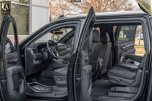 Image of a black armored GMC Yukon Denali XL SUV with its doors open showing a view of the interior, including the front and rear seats, dashboard, and steering wheel. The vehicle is parked outside near a building with large windows.