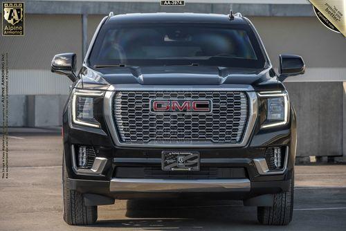 Front view of a black arnmored GMC Yukon Denali SUV with dual exhaust pipes, displayed in a parking area. The vehicle has "Alpine Armoring" branding on the back.