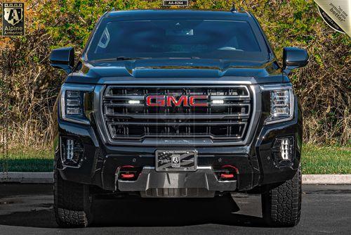 A black GMC Yukon XL AT4 truck is parked facing forward on a paved surface with greenery and a building in the background.