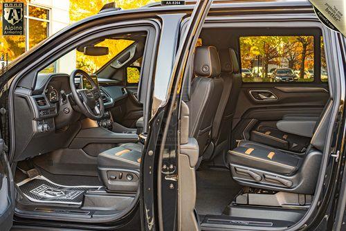 Interior view of a black armored GMC Yukon XL AT4 SUV showcasing the driver and front passenger seats, as well as the second-row captain's chairs, with leather upholstery and yellow stitching. The doors are open.