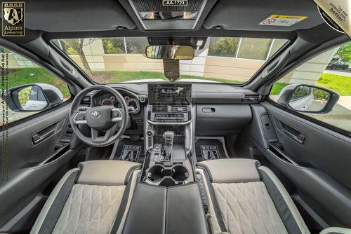 Interior view of an Armored Toyota Land Cruiser 300 showing the steering wheel, dashboard, touchscreen, and leather seats. Visible logos include Alpine Armoring on the left and top of the image.