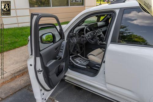 The driver-side door of an Armored Toyota Land Cruiser 300 is open, showing the interior with a steering wheel, dashboard, and seat. The car is parked in a lot next to a sidewalk and a building.