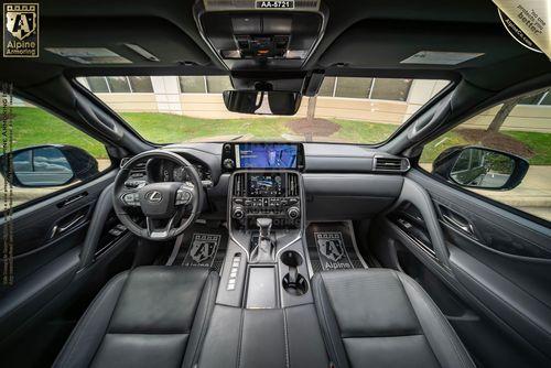 Interior view of a Lexus LX600 Luxury sedan with leather seats and a modern dashboard featuring a large central touchscreen display and various controls. The steering wheel prominently displays the Lexus logo.