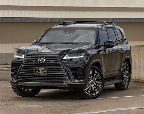 A black armored Lexus LX600 Luxury SUV is parked in an outdoor setting with a concrete wall in the background. The vehicle features a prominent front grille and sleek headlights.