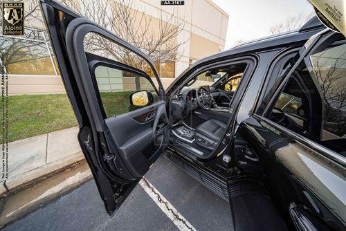 An open driver's side door of a black Lexus LX 600 Ultra Luxury SUV revealing the interior, including the steering wheel, dashboard, and front seats. The vehicle is parked near a sidewalk with some trees and a building visible.