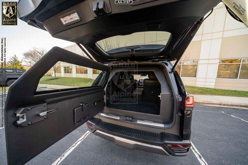 A Lexus LX 600 Ultra Luxury with its rear hatch open, revealing an armored door and an additional compartment inside. The scene is set in a parking lot beside a modern building.