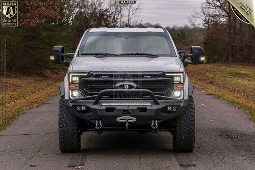 A white Mastiff, modified from a Ford Super Duty truck, with aftermarket bumper and winch is parked on a rural road, surrounded by trees.