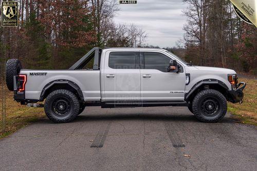 Side view of a white off-road pickup Mastiff  with black trims and rugged tires is parked on a road surrounded by trees. It features a mounted spare tire on the back and a roll bar over the bed.